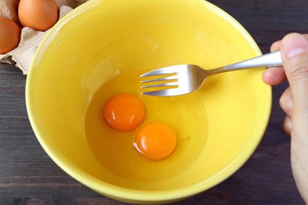 Raw eggs in a yellow bowl about to be whisked by a hand holding a fork.