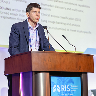 Scott Palmer speaks at a podium presenting on respiratory research at a conference, with a screen behind displaying study capabilities and a RIS 2026 logo.
