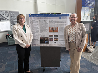 Two presenters stand on either side of a research poster displayed on a board in a conference setting; the poster includes text, charts, and images, and exhibits are visible in the background.