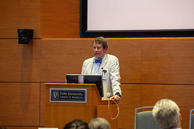 A speaker stands behind a podium with a “Duke University School of Medicine” sign, presenting in front of a projection screen in a lecture hall-style room.