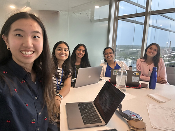 Five STAR program participants sit around a conference table with open laptops during a meeting. The group faces the camera in a bright room with large windows overlooking the campus.