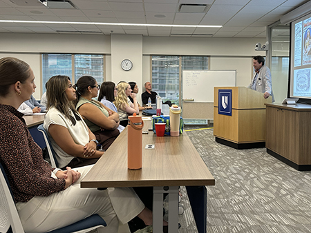 Participants seated around conference tables listen to Robert Califf speaking at a podium in a classroom-style meeting room. A projected presentation is displayed on a screen at the front of the room.