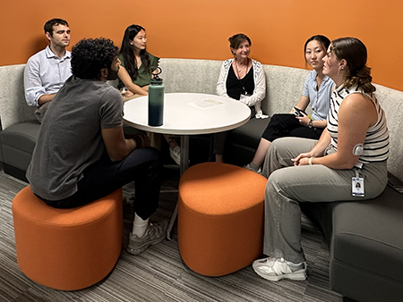 Six STAR Program participants sit together in a circular seating area, discussing ideas around a round table. Orange stools and an orange accent wall create an informal meeting space designed for collaboration and conversation.