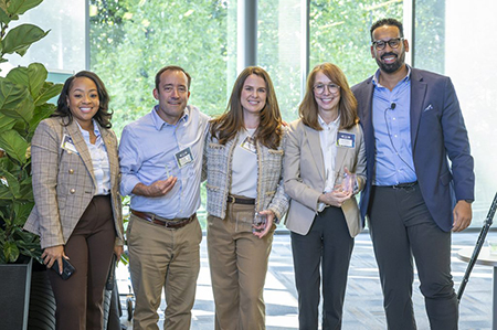 Cross Campus Catalyst award recipients pose together indoors, each holding a glass award, in front of large windows and greenery.