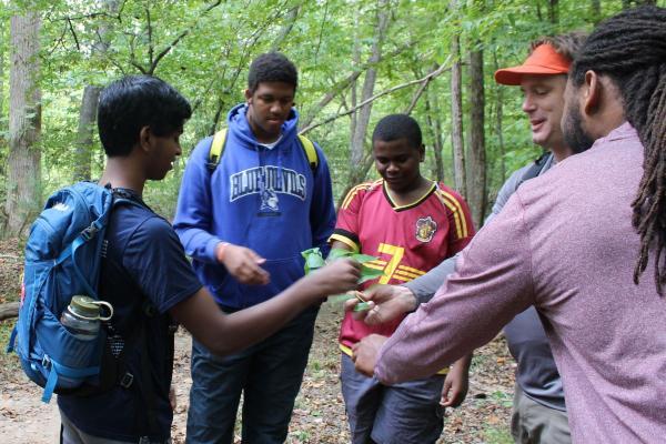 Three teens are seen smiling and interacting with two adult guides in a forest.