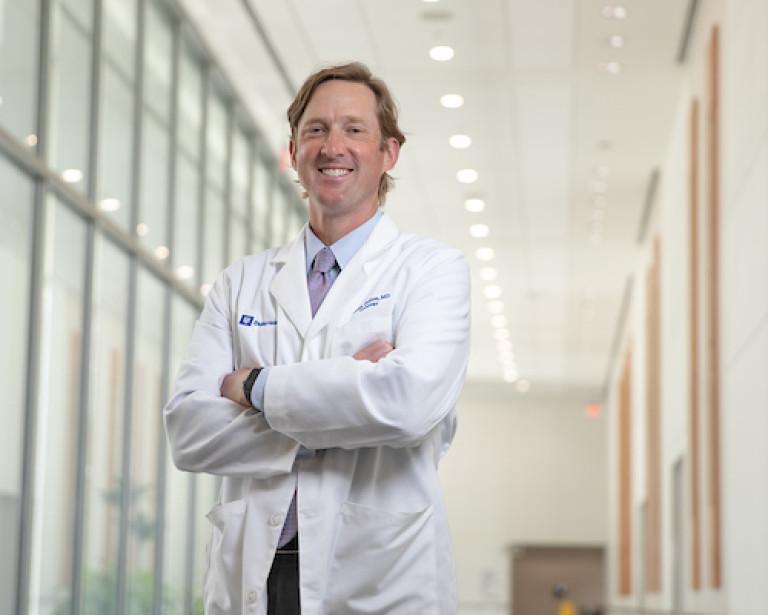 DCRI faculty member Adam DeVore stands in his white coat, smiling, in the midst of a hallway at Duke Health.