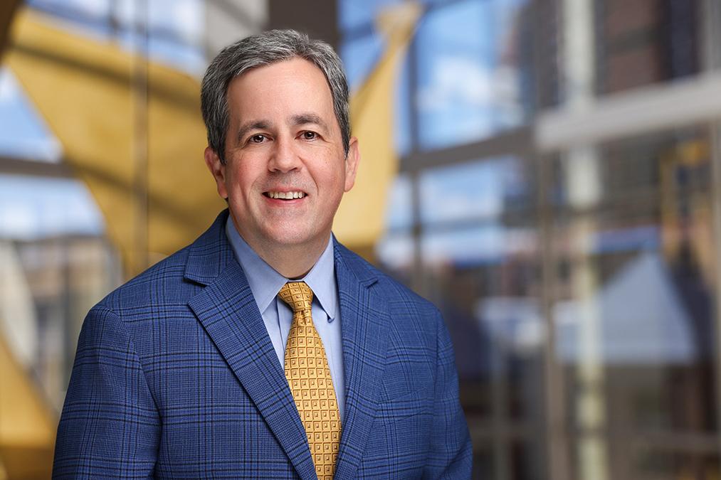 DCRI's Adrian Hernandez is shown smiling in a blue suit jacket, blue shirt and yellow tie.