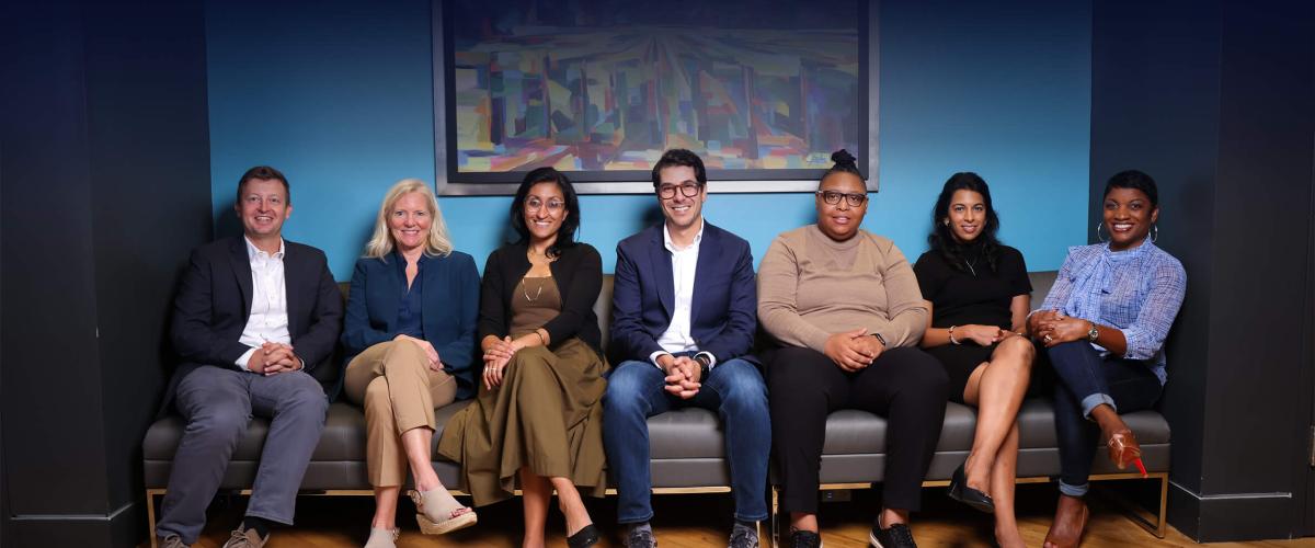 The team of seven i-Cubed leaders sit on a couch, smiling against a deep blue background.