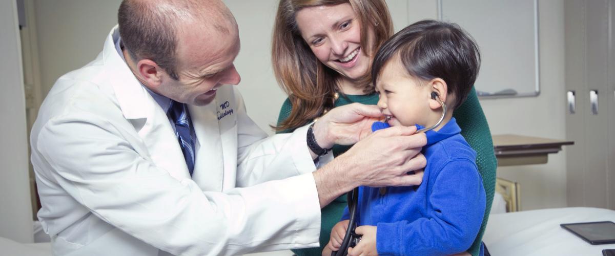 A doctor gently allows his pediatric patient to listen to the stethoscope, as the child laughs and sits and on his smiling mother's lap.