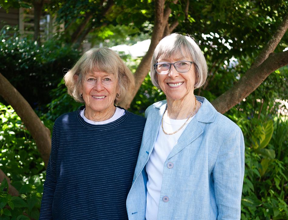 Two women stand together in front of a tree. 