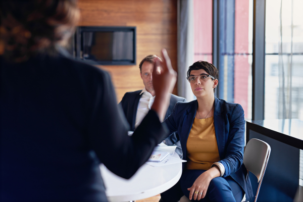 Two people sit in a meeting listening attentively to a third person whose back is turned to the viewer.