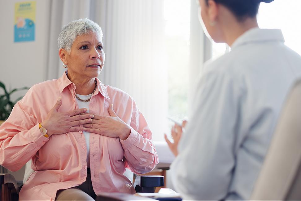 A woman speaks to a healthcare provider in a white coat while gesturing to her heart. 