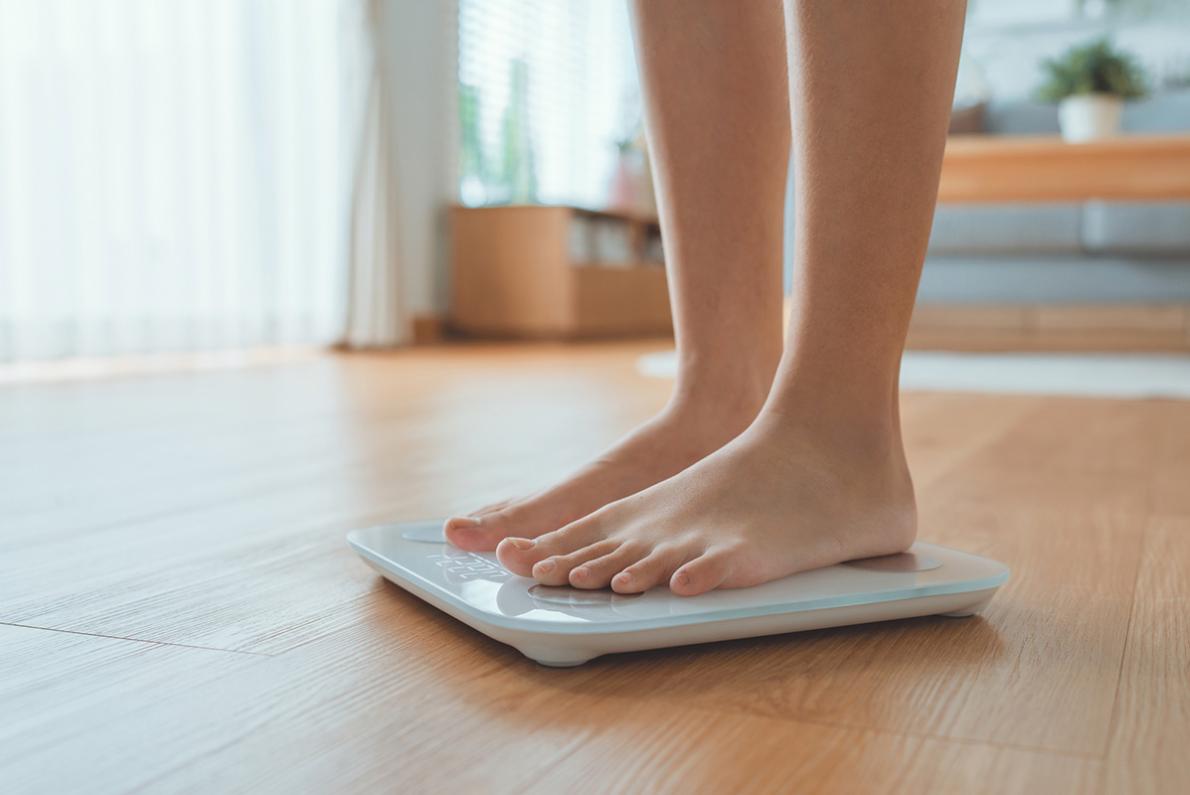 A woman's feet are seen standing on a white scale sat on a hardwood floor as she measures her weight.