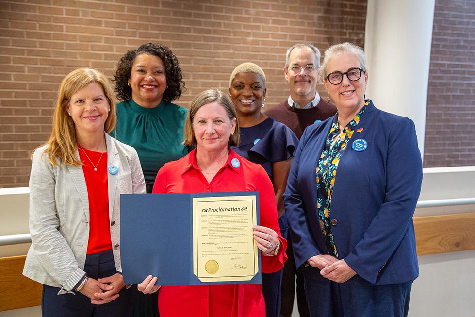 Six individuals stand smiling. The person in the center is holding a blue folder containing a paper labeled "Proclamation" at the top, with a gold seal at the bottom.